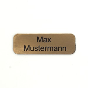 Puede incluir: Una etiqueta de nombre de metal de color dorado con el texto "Max Mustermann" grabado en negro.