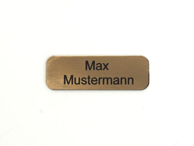 Puede incluir: Una etiqueta de nombre de metal de color dorado con el texto "Max Mustermann" grabado en negro.