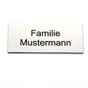 K&ouml;nnte beinhalten: Eine silberne Namensschilder mit schwarzem Text, der "Familie Mustermann" lautet.