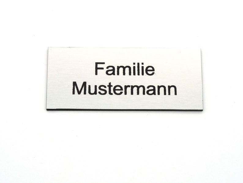 K&ouml;nnte beinhalten: Eine silberne Namensschilder mit schwarzem Text, der "Familie Mustermann" lautet.