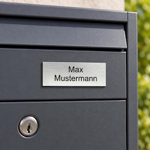 Könnte beinhalten: Ein dunkelgrauer Briefkasten aus Metall mit einem rechteckigen, gebürsteten silbernen Namensschild in der Mitte der Frontplatte. Das Namensschild zeigt den schwarzen Aufdruck Max Mustermann. Ein silbernes Rundschloss befindet sich darunter.
