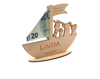 Könnte beinhalten: Ein hölzerner Geschenkhalter in Bootsform mit einem Segel aus einem 20-Euro-Schein. Das Boot zeigt ausgeschnittene Tierfiguren und den Namen "LINDA" mit dem Datum 10.02.2024. Das Segel trägt den Text "ZUR TAUFE ALLES GUTE".