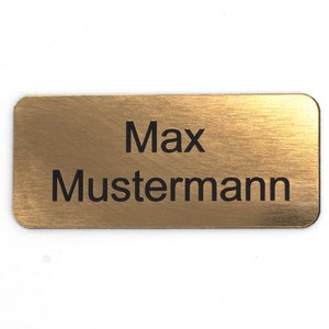 Puede incluir: Una etiqueta de nombre de metal de color dorado con el texto "Max Mustermann" grabado en negro.