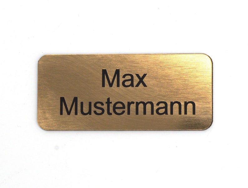 Puede incluir: Una etiqueta de nombre de metal de color dorado con el texto "Max Mustermann" grabado en negro.