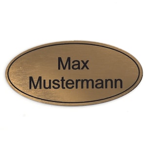 Puede incluir: Una etiqueta de nombre ovalada dorada con un borde negro y el texto "Max Mustermann" grabado en ella.