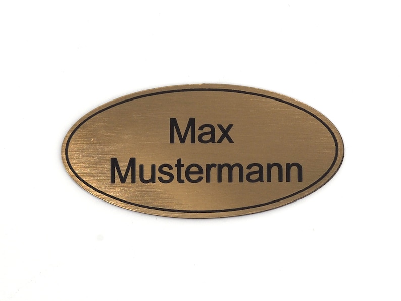 Puede incluir: Una etiqueta de nombre ovalada dorada con un borde negro y el texto "Max Mustermann" grabado en ella.