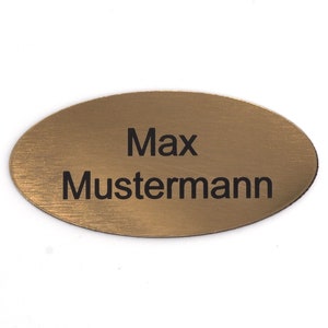 Puede incluir: Una etiqueta de nombre ovalada dorada con el texto "Max Mustermann" grabado en negro.