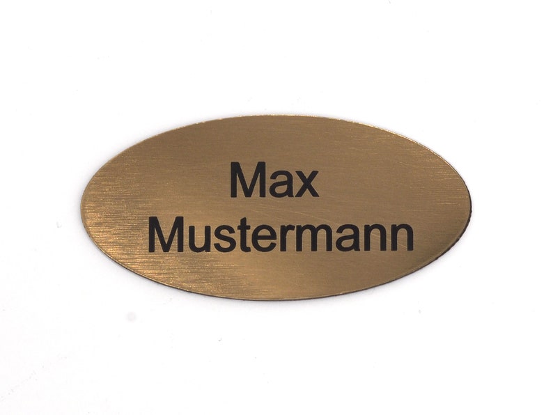 Puede incluir: Una etiqueta de nombre ovalada dorada con el texto "Max Mustermann" grabado en negro.