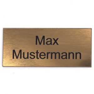 Puede incluir: Una placa de nombre de metal de color dorado con el texto "Max Mustermann" grabado en negro.