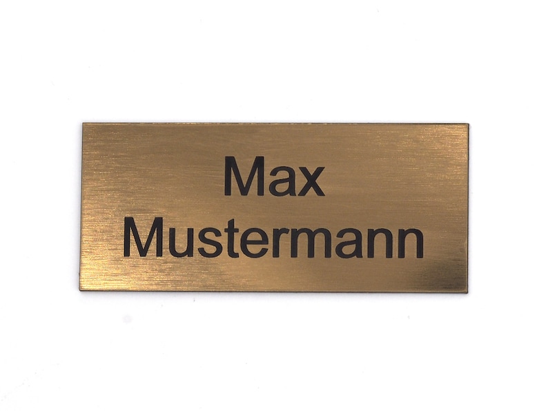 Puede incluir: Una placa de nombre de metal de color dorado con el texto "Max Mustermann" grabado en negro.
