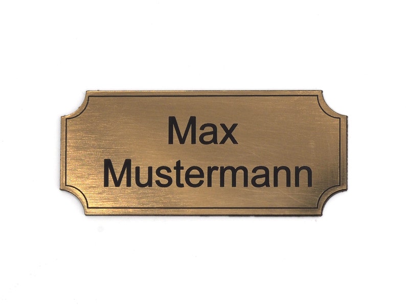 Puede incluir: Una placa de nombre de metal de color dorado con esquinas redondeadas y acabado cepillado. La placa tiene el texto "Max Mustermann" grabado en negro.