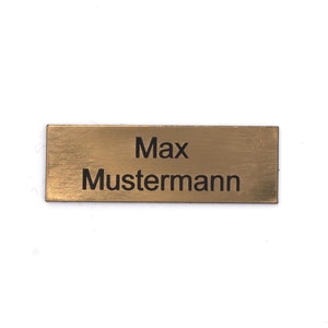 Puede incluir: Una placa de nombre de metal de color dorado con el texto "Max Mustermann" grabado en negro.