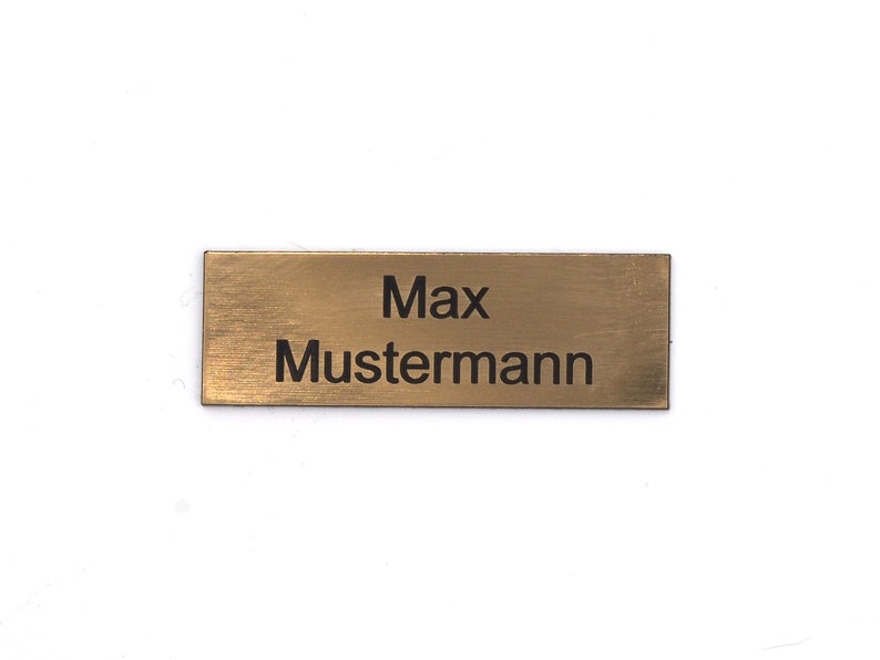 Puede incluir: Una placa de nombre de metal de color dorado con el texto "Max Mustermann" grabado en negro.
