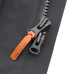 Könnte beinhalten: Ein schwarzer Reißverschluss mit einem braunen Leder-Zipper. Der Zipper ist mit dem Text "Sophia 0123 4567 890" graviert.