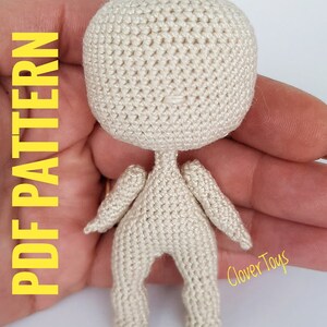May include: A crocheted doll pattern in the form of a small, beige, crocheted doll with arms and legs. The doll is about 4 inches tall and is being held in a person's hand. The text "Clover Toys" is visible in the bottom right corner of the image.