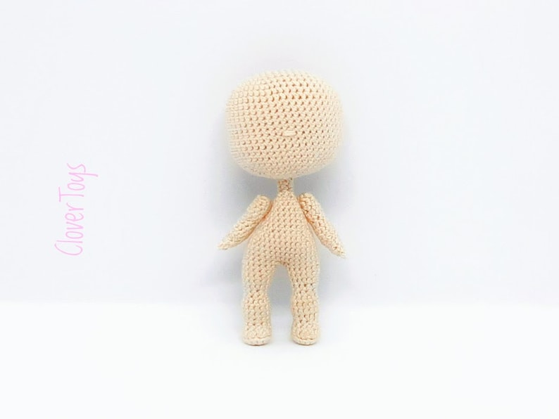 May include: A crocheted doll with a beige body and no facial features. The doll is standing on a white surface.