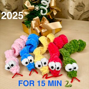 May include: Five crocheted snake toys in different colours: pink, blue, yellow, red, and green. Each snake has a white crocheted eye and a black pupil. The text "FOR 15 MIN" is written below the snakes. The year "2025" is written in the top left corner of the image.
