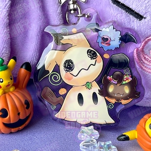 May include: A clear acrylic keychain featuring a cartoon ghost Pokemon with a black and white face, holding a trick-or-treat bag. The ghost is wearing a green bug on its shoulder. The background is purple with a swirl pattern and a small blue bat Pokemon.