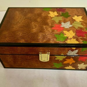 May include: A wooden box with a hinged lid and a brass latch. The box is decorated with a design of colourful autumn leaves.