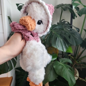 May include: A handmade plush goose toy with a white body, orange beak and feet, and a pink hat and scarf. The goose has black eyes and is made of soft, fluffy material. The toy is held up in front of a background of green plants.