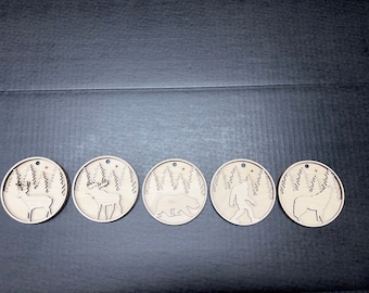 May include: Five wooden ornaments with a winter forest theme. Each ornament features a different animal silhouette: a deer, a moose, a bear, a Bigfoot, and a wolf. The ornaments are all circular and have a rustic, natural look.