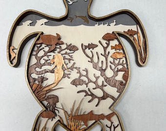 May include: A wooden turtle sculpture with a layered design. The turtle's shell is cut out to reveal a scene of a mermaid swimming in a coral reef with fish and seagrass. The top layer of the sculpture shows a sun and birds flying in the sky.