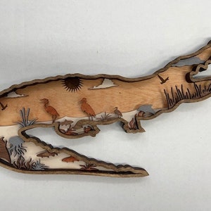 May include: A wooden alligator cutout with a detailed design of birds, fish, and plants. The alligator is brown and the design is in shades of brown, white, and blue.