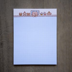 Peut inclure: Un bloc-notes rectangulaire avec un quadrillage bleu clair. La bordure supérieure présente une ligne horizontale avec une rangée de petits animaux de dessin animé colorés, dont un ours central dans un lit bleu, avec le texte minch imprimé verticalement.
