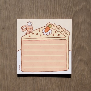 May include: A square memo pad featuring a light orange illustration of a layered carrot cake slice. A small brown rabbit wearing a white chef hat stands on the top left corner. A small orange carrot sits on white frosting. The word minch is printed.