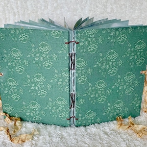 May include: A small, green fabric-covered journal with a floral pattern. The journal is closed and has brown ribbon ties.