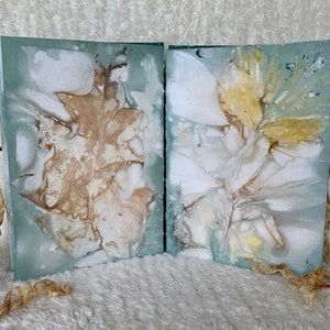 May include: Two handmade journals with pressed leaves and flowers on the covers. The covers are a light blue colour with a white background. The journals are tied together with a brown ribbon.