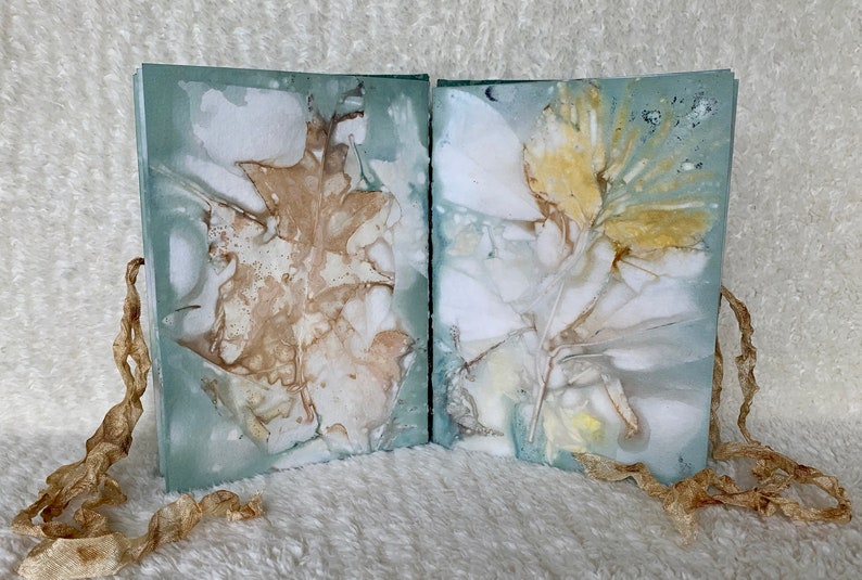 May include: Two handmade journals with pressed leaves and flowers on the covers. The covers are a light blue colour with a white background. The journals are tied together with a brown ribbon.