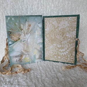May include: A handmade journal with a green cover and a white floral design. The journal has a white ribbon closure and the words "Handmade with Love" are written on the front cover.