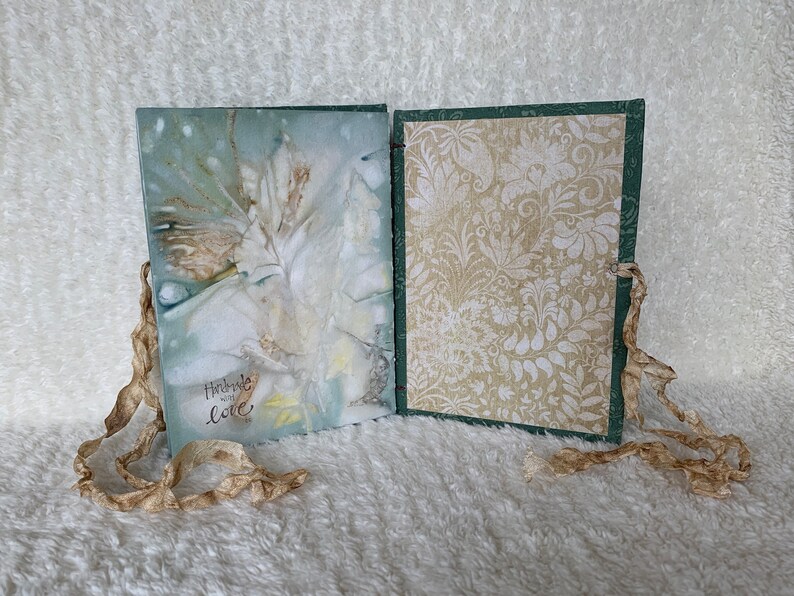 May include: A handmade journal with a green cover and a white floral design. The journal has a white ribbon closure and the words "Handmade with Love" are written on the front cover.