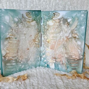 May include: An open book with a blue-green cover and white pages. The pages are decorated with abstract patterns in shades of white, brown, and pink. The book is lying on a white fluffy surface with two pieces of brown ribbon on either side.