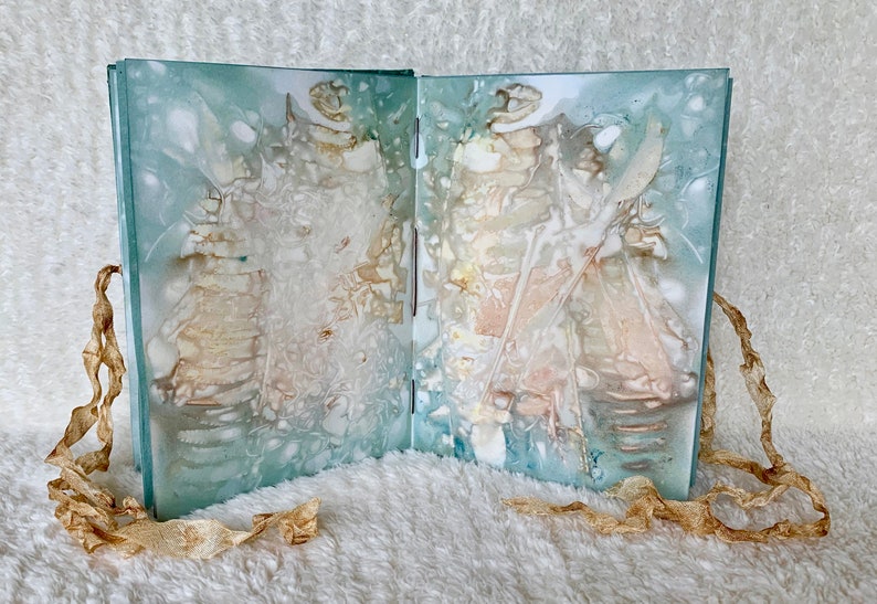 May include: An open book with a blue-green cover and white pages. The pages are decorated with abstract patterns in shades of white, brown, and pink. The book is lying on a white fluffy surface with two pieces of brown ribbon on either side.