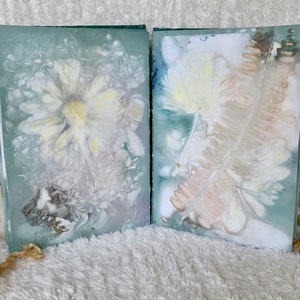 May include: Two pages of a handmade book featuring pressed flowers and leaves. The pages are a pale blue-green colour and the flowers are white and yellow. The book is bound with a brown ribbon.