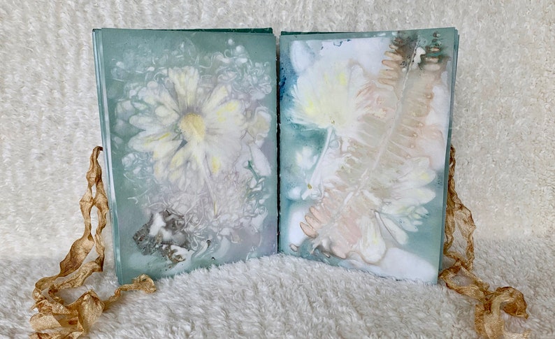 May include: Two pages of a handmade book featuring pressed flowers and leaves. The pages are a pale blue-green colour and the flowers are white and yellow. The book is bound with a brown ribbon.