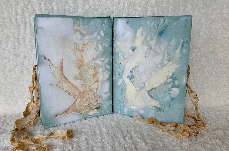 May include: A handmade book with a blue and white marbled cover. The book is open to reveal two pages with pressed leaves and flowers. The book is tied together with a brown ribbon.