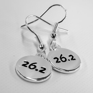 May include: Silver earrings with a hook design. Each earring has a small, oval charm with the number "26.2" in black, representing a marathon distance. The earrings are shown against a white background.