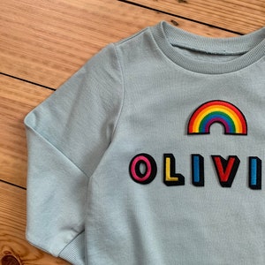May include: A light blue sweatshirt with a rainbow patch and the name "OLIVI" spelled out in colourful letters.