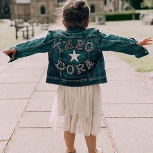 May include: A child wearing a blue denim jacket with the name "Theo Dora" and a white star on the back. The child is wearing a white tulle skirt and is standing on a pavement.