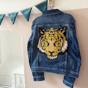 May include: A blue denim jacket with an embroidered tiger face on the back, in shades of yellow and brown.