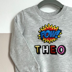 May include: Grey sweatshirt with a colourful "POW!" graphic and the name "THEO" in colourful letters.