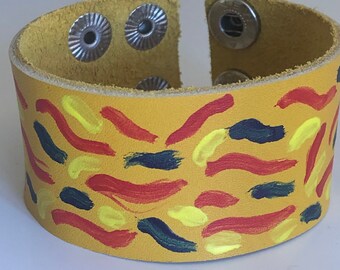 Op de afbeelding: Een gele leren armband met een kleurrijk abstract ontwerp dat erop is geschilderd. De armband heeft drie zilveren drukknopen.