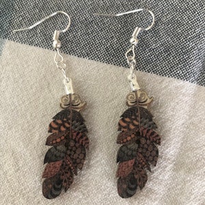 Puede incluir: Un par de pendientes de aro de plata con un diseño de pluma marrón y negro. Las plumas tienen un patrón detallado y están unidas a los pendientes con un aro de plata.