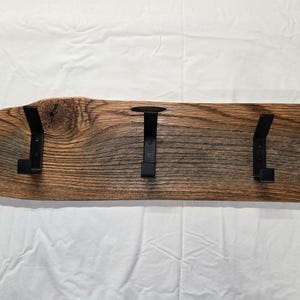 Puede incluir: Un perchero de madera rústica con cinco ganchos de metal negro. La madera tiene un aspecto natural y envejecido con vetas y nudos visibles. Los ganchos están espaciados uniformemente a lo largo de la tabla, midiendo unos 90 cm de largo.
