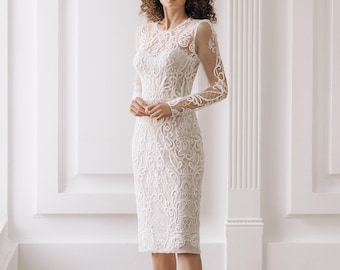 May include: A white, knee-length dress with detailed lace patterns. It has long, sheer sleeves and a high neckline. The model wears silver high heels. The background is a white wall with architectural moulding.