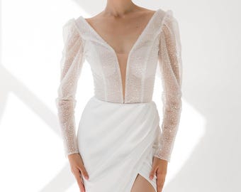 Op de afbeelding: Een witte trouwjurk met een diepe V-hals en lange, doorschijnende mouwen versierd met kleine, glinsterende details. De jurk heeft een getailleerd lijfje en een zwierige rok met een hoge split, wat een elegante uitstraling creëert. Geschikt voor een formele gelegenheid.