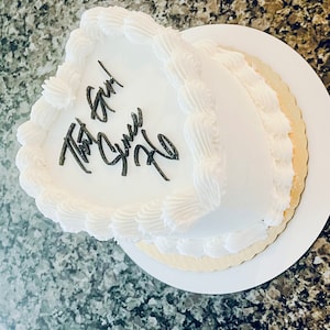 May include: A white two-tiered cake with white frosting and a heart-shaped top. The cake is decorated with a simple black script that says "The Girl Who"